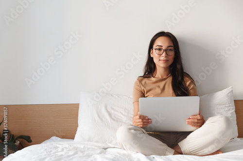 Portrait of attractive young asian woman in glasses, sit on bed and using laptop, smiling at camera. Girl working from home with computer. Female student prepare homework online