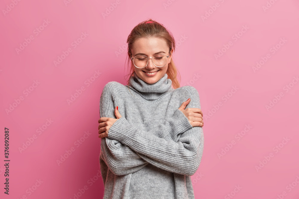 Happy tender girl dressed in grey warm oversized sweater, embraces herself, enjoys comfort during rainy autumn day, wears transparent glasses smiles pleasantly, isolated over pink background