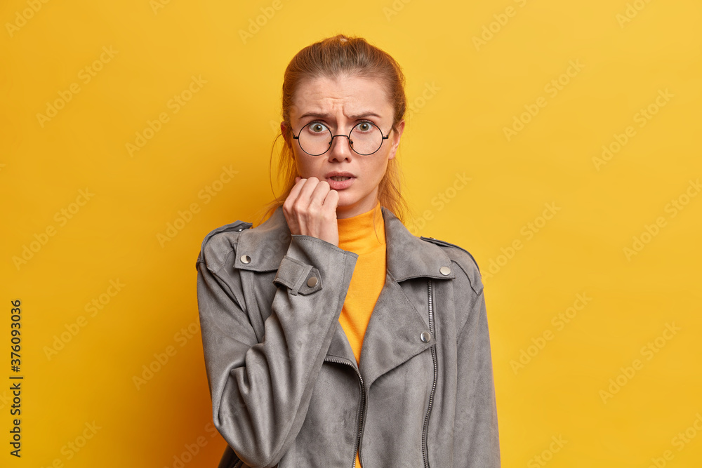 Photo of shocked worried woman astonished with bad concerning news, looks nervously at camera, stands anxious, dressed in grey jacket, poses against yellow background, being alarmed and insecure Photo of shocked worried woman astonished with bad concerning news, looks nervously at camera, stands anxious, dressed in grey jacket, poses against yellow background, being alarmed and insecure