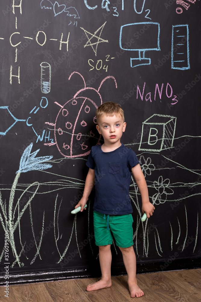 three year old boy draws with colorful chalk on a large blackboard. black wall with children's drawings and formulas. vertical photography. look into the camera.