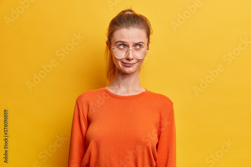 Photo of thoughtful good looking nerdy girl in spectacles, has thoughtful expression, concentrated at something, dressed in orange jumper, isolated on yellow background, witnesses terrible crime
