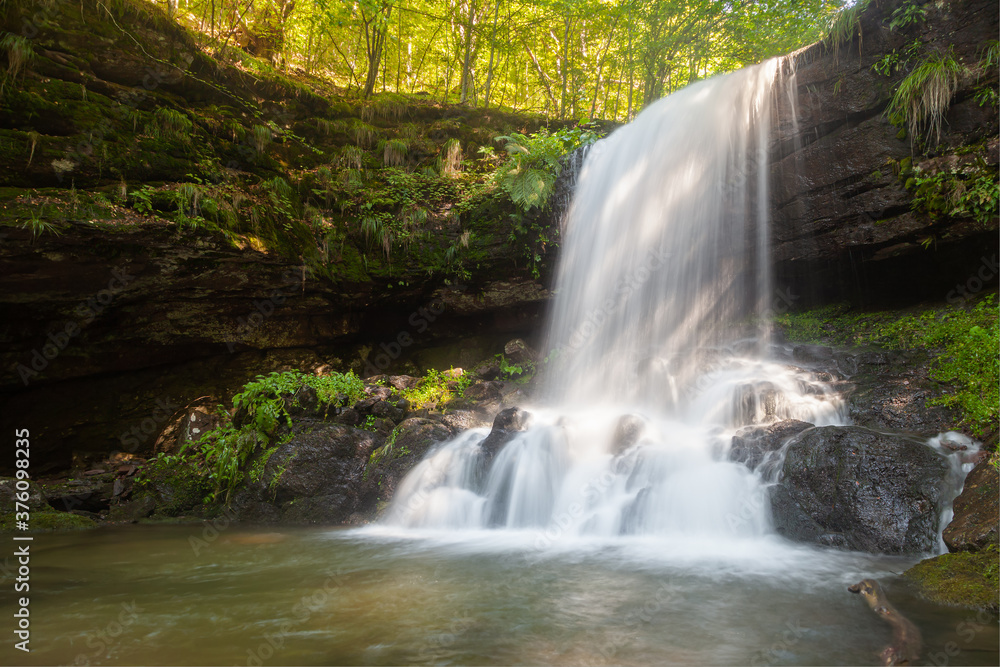 Obraz premium Amazing sunrays on scenic Skok waterfall, long exposure blurred motion of the water and vivid green plants on the wet, dark rocks