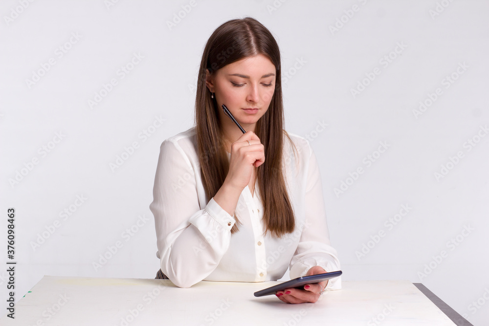 A young business woman a background isolated in white, in white shirts, sitting at a table, writing with a stylus on a tablet A young business woman a background isolated in white, in white shirts, sitting at a table, writing with a stylus on a tablet