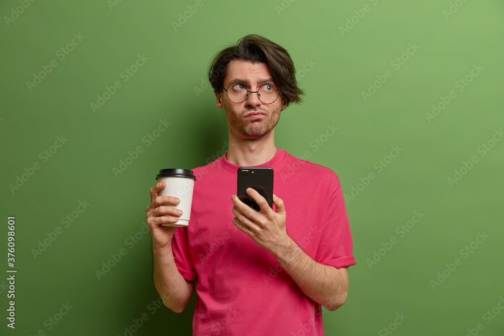 Serious adult guy with pensive expression, has bristle, holds mobile phone, surfs internet, sends text messages, drinks coffee to go, wears pink t shirt, stands against vivid green background Serious adult guy with pensive expression, has bristle, holds mobile phone, surfs internet, sends text messages, drinks coffee to go, wears pink t shirt, stands against vivid green background