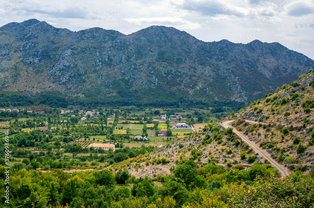 View of valley and small village in Albanian mountains. Summer landscape with green trees, road on a hill and clouds on the sky. 