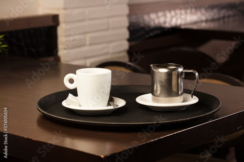 cup of freshly brewed hot coffee with metal milk container, on the waiter's tray, on the wooden table, inside the cafeteria and free space for decoration.