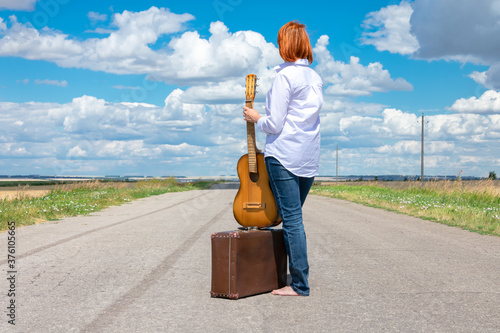 A young girl with red hair and a white shirt stands barefoot on the road, holding a guitar and an old suitcase at her feet. The girl is hitchhiking. photo