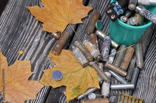 Corroded used batteries. They lie in and around the trash can. Dried maple leaves lie nearby. Disposal of hazardous waste.