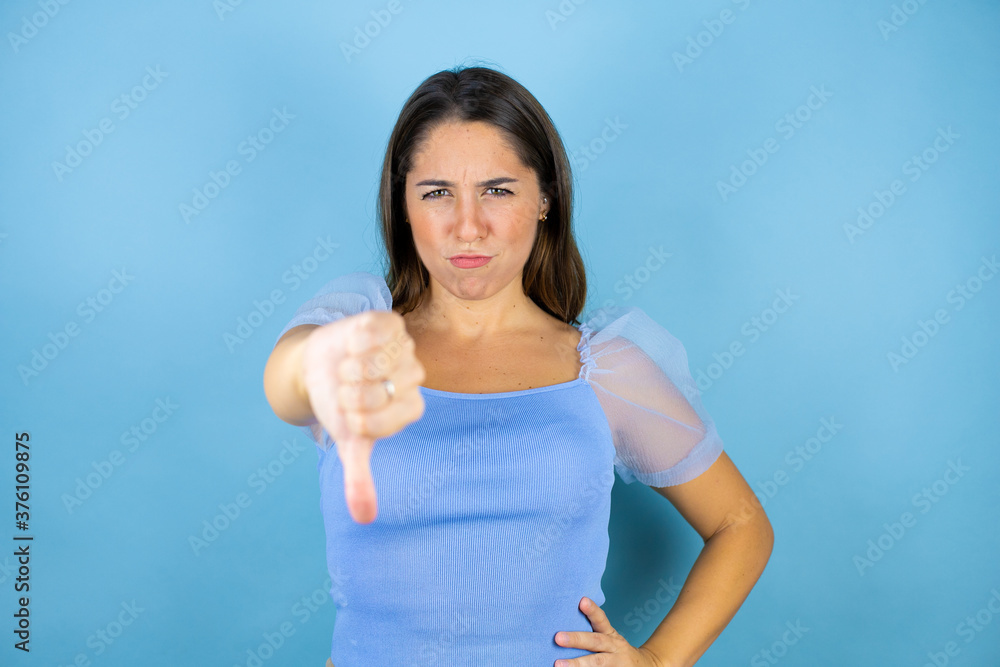 Young beautiful woman over isolated blue background with angry face, negative sign showing dislike with thumb down Young beautiful woman over isolated blue background with angry face, negative sign showing dislike with thumb down