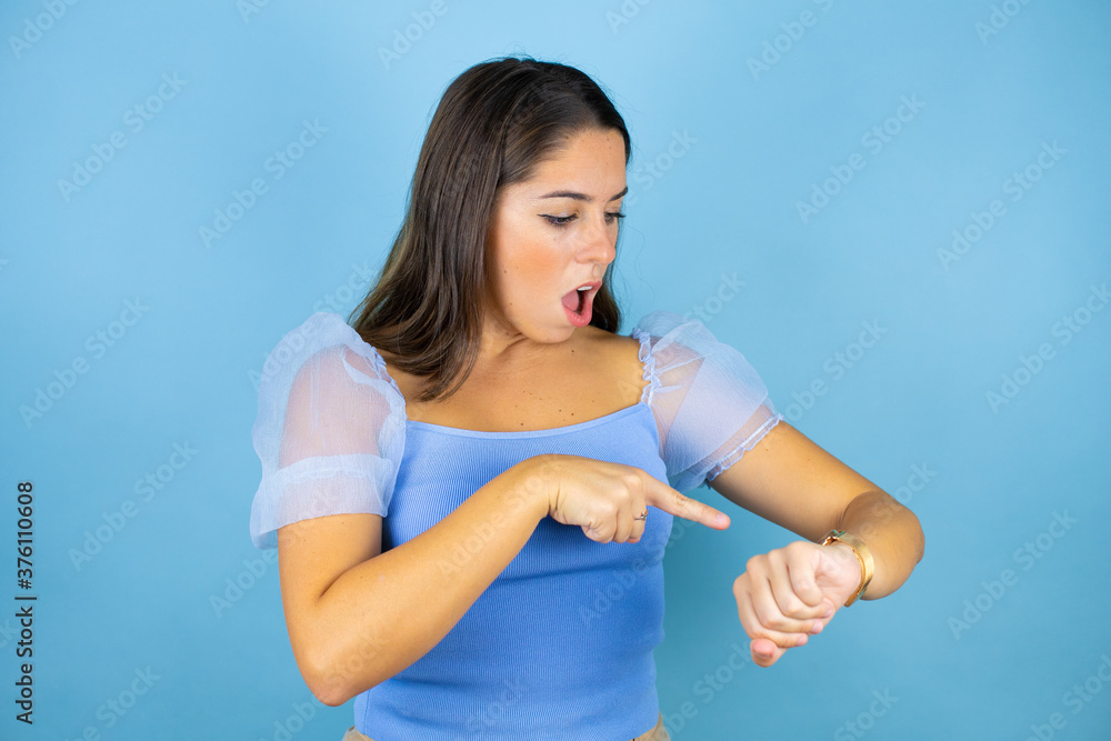 Young beautiful woman over isolated blue background surprised and pointing his watch because it's late Young beautiful woman over isolated blue background surprised and pointing his watch because it's late