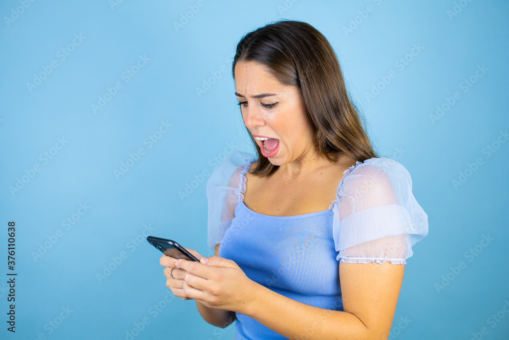 Young beautiful woman over isolated blue background chatting with her phone and scared Young beautiful woman over isolated blue background chatting with her phone and scared