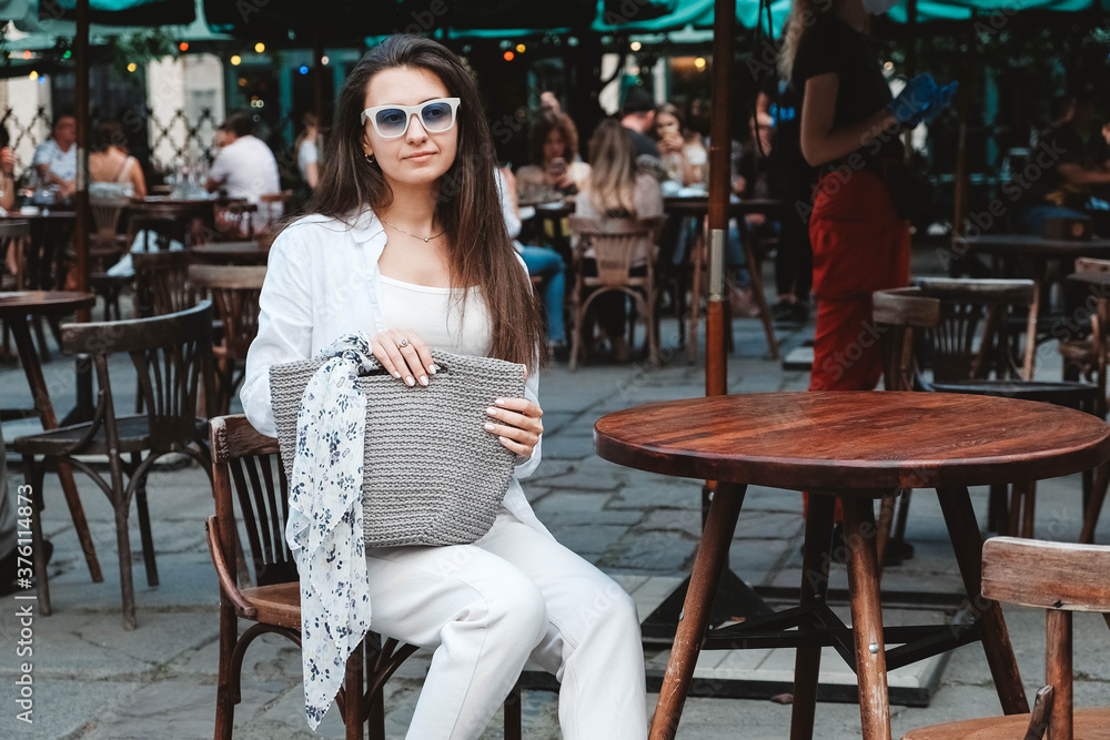Beautiful young brunette woman wearing in sunglasses and white clothes, holding knitted bag sitting at the table of a street cafe. Copy, empty space for text Beautiful young brunette woman wearing in sunglasses and white clothes, holding knitted bag sitting at the table of a street cafe. Copy, empty space for text