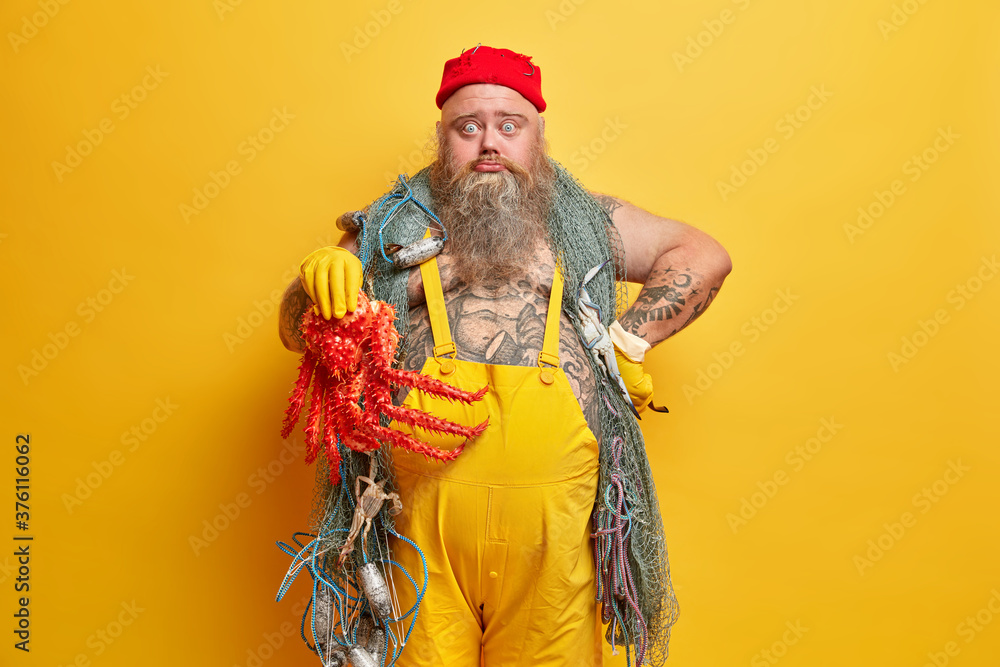Obese bearded male sailor surprised to catch octopus instead of fish, looks with shocked sad expression, has summer vacation, poses against yellow background. Member of ship crew. Yacht voyage