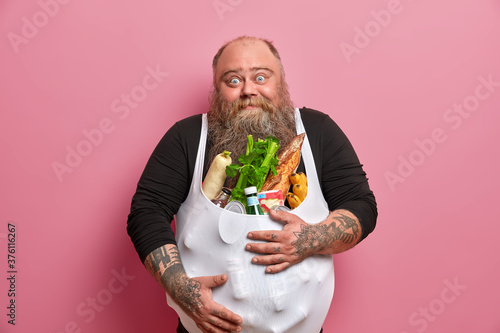 Obese bearded European man has excess weight because of unhealthy harmful nutrition, carries different products, poses against pink background, covers abdomen with food, fights against obesity
