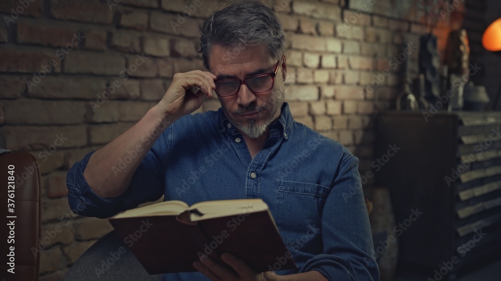 Mature man in his 50s reading book at home sitting in dark living room. Mature man in his 50s reading book at home sitting in dark living room.