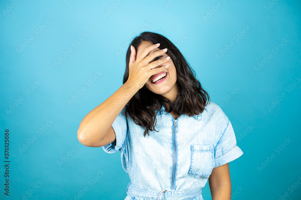 Young beautiful woman wearing a denim jumpsuit over isolated blue background peeking in shock covering face and eyes with hand, looking through fingers with embarrassed expression Young beautiful woman wearing a denim jumpsuit over isolated blue background peeking in shock covering face and eyes with hand, looking through fingers with embarrassed expression