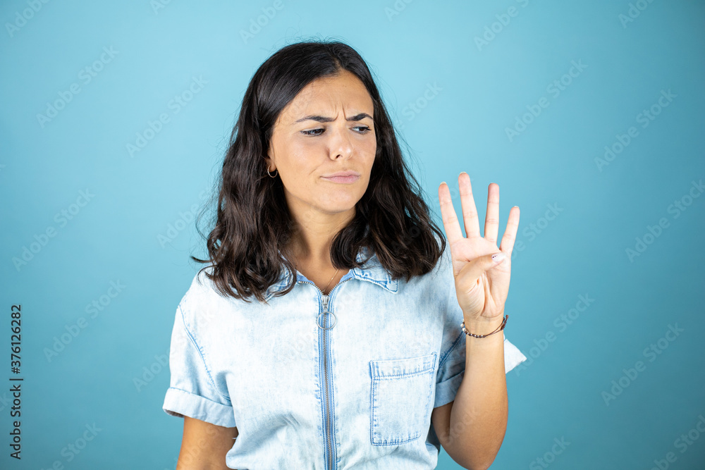 Young beautiful woman wearing a denim jumpsuit over isolated blue background showing and pointing up with fingers number four while smiling confident and happy. Young beautiful woman wearing a denim jumpsuit over isolated blue background showing and pointing up with fingers number four while smiling confident and happy.