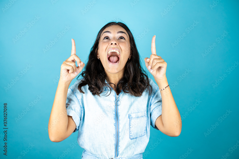 Young beautiful woman wearing a denim jumpsuit over isolated blue background amazed and surprised looking and pointing up with fingers and raised arms. Young beautiful woman wearing a denim jumpsuit over isolated blue background amazed and surprised looking and pointing up with fingers and raised arms.