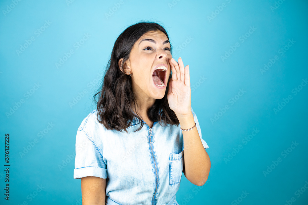 Young beautiful woman wearing a denim jumpsuit over isolated blue background shouting and screaming loud to side with hands on mouth. Young beautiful woman wearing a denim jumpsuit over isolated blue background shouting and screaming loud to side with hands on mouth.
