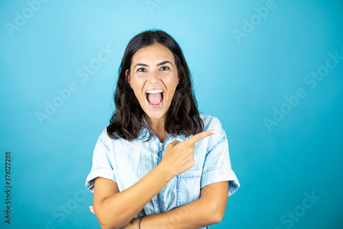 Young beautiful woman wearing a denim jumpsuit over isolated blue background smiling and pointing with hand and finger to the side