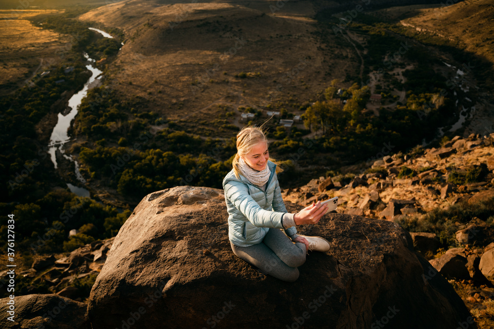 Young caucasian female sitting on lush mountain top taking selfie with smartphone at sunset.