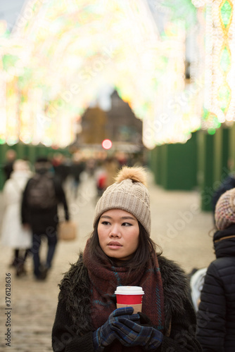 Asian girl wearing a woolly hat, scarf and gloves holding a warm cup of coffee as she walks and looks away in front of a light display in George Street, Edinburgh City Center, UK.