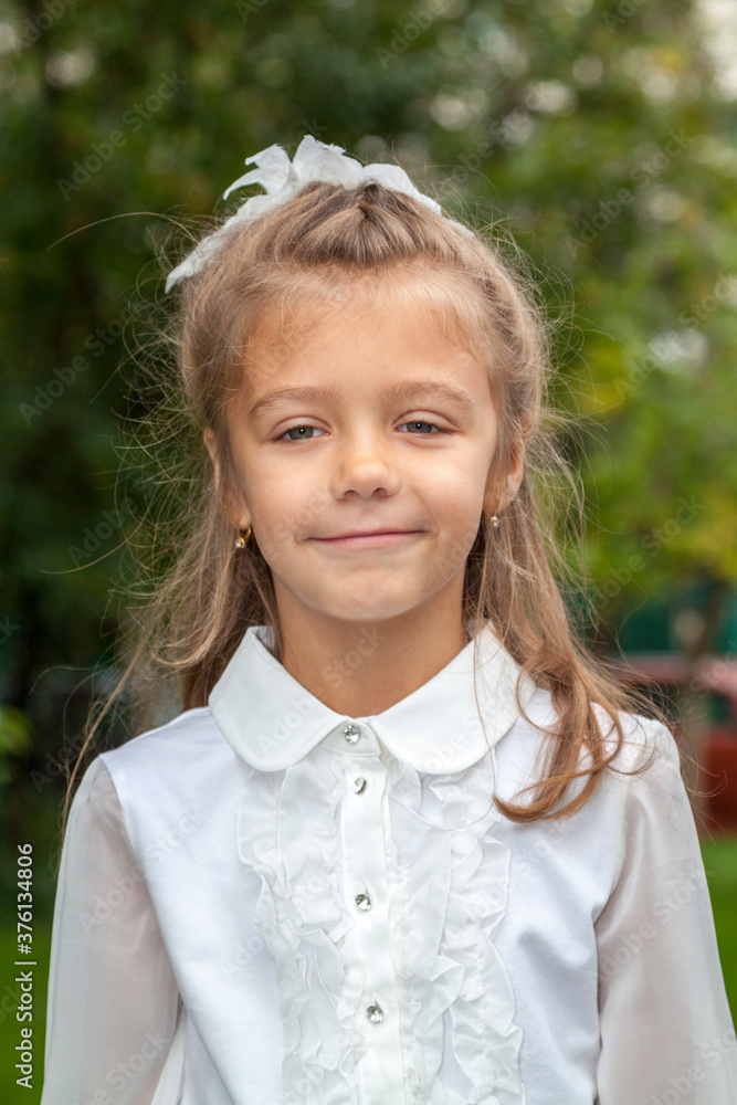 Portrait of cute smiling little caucasian girl in white blouse and bow, against green of summer park. Child go to school. Portrait of cute smiling little caucasian girl in white blouse and bow, against green of summer park. Child go to school.