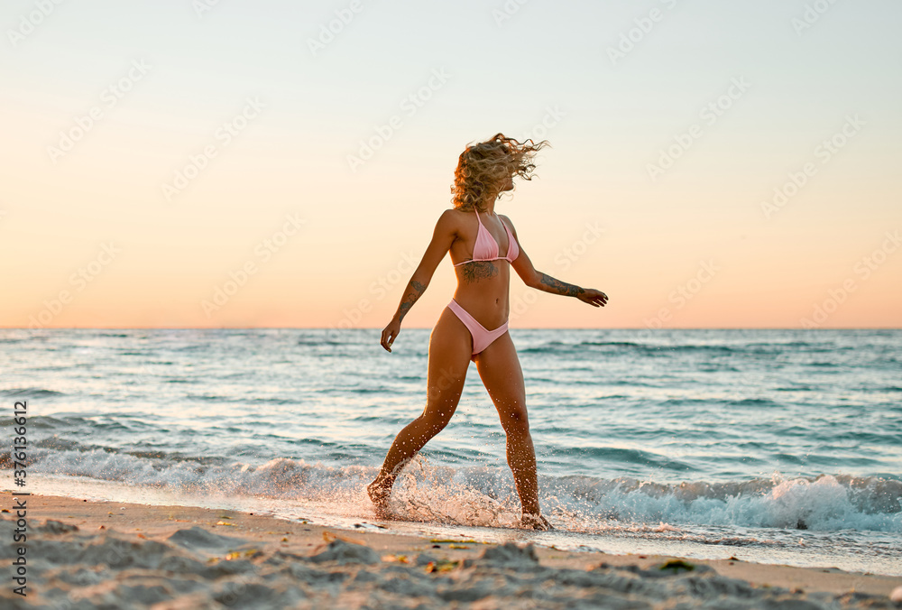 Naklejka premium Attractive Caucasian woman in bikini swimsuit strolls on the beach at sunrise enjoying the sound of the waves.