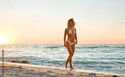 Attractive Caucasian woman in bikini swimsuit strolls on the beach at sunrise enjoying the sound of the waves. © Valerii Apetroaiei