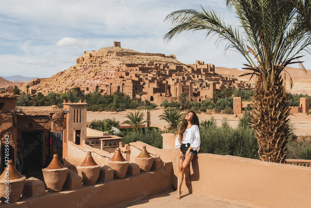 Beautiful young woman happy to travel in Morocco. Ait-Ben-Haddou kasbah on background. Wearing in white shirt and jeans shorts. - obrazy, fototapety, plakaty 