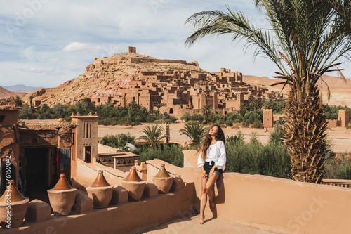 Beautiful young woman happy to travel in Morocco. Ait-Ben-Haddou kasbah on background. Wearing in white shirt and jeans shorts.