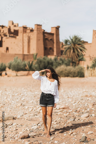 Young curly woman eastern appearance walking on background of kasbah Ait-Ben-Haddou. Travel in Morocco, Ouarzazate. Summer vacations, travel lifestyle concept. photo