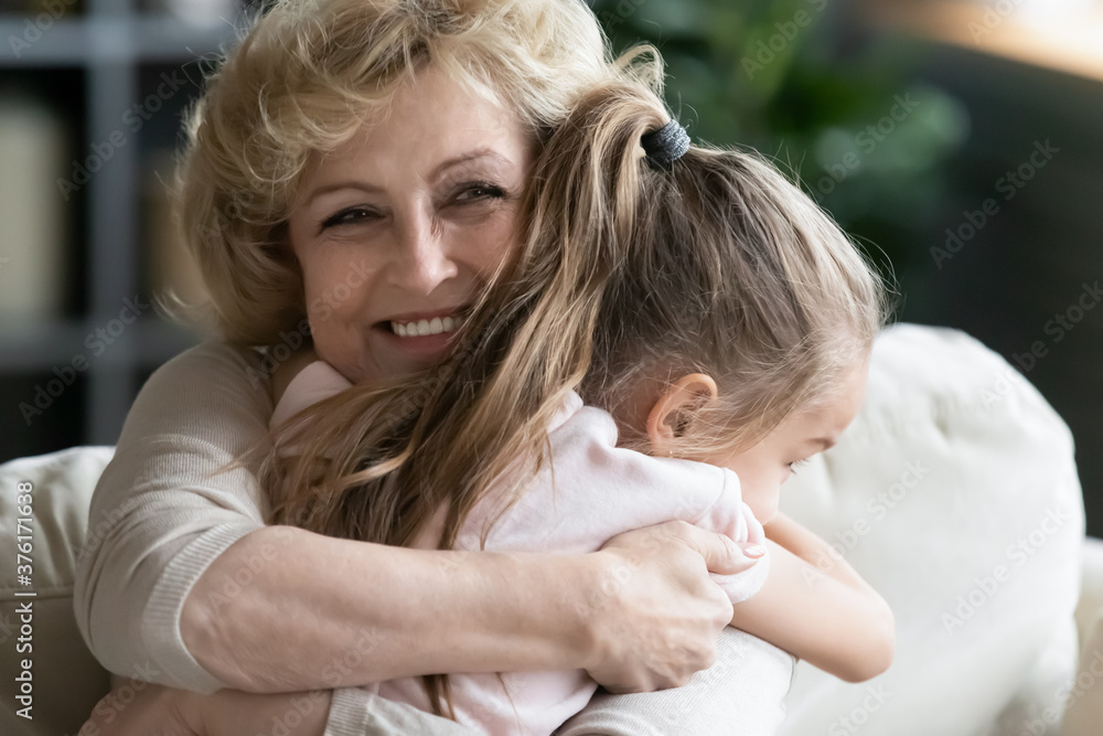 Close up smiling loving mature woman grandma hugging little granddaughter, happy grandmother and pretty girl enjoying tender moment, cuddling, spending leisure time together at home
