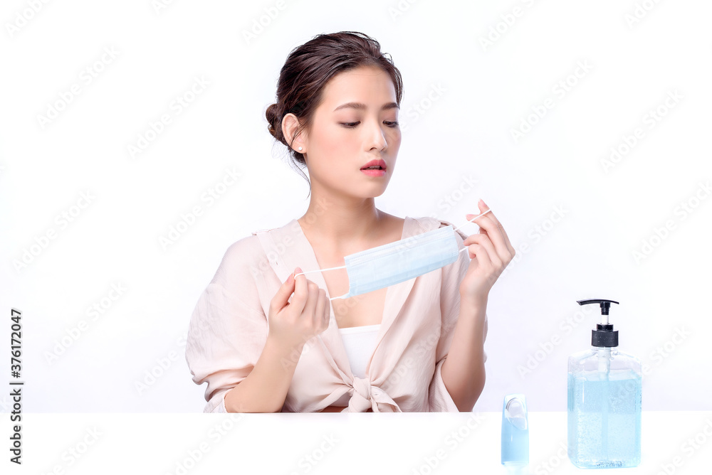 Portrait of beautiful charming young Asian woman posing with surgical mask or face mask to protect from corona virus and sitting with white table on white background, health care protection Covid-19 Portrait of beautiful charming young Asian woman posing with surgical mask or face mask to protect from corona virus and sitting with white table on white background, health care protection Covid-19