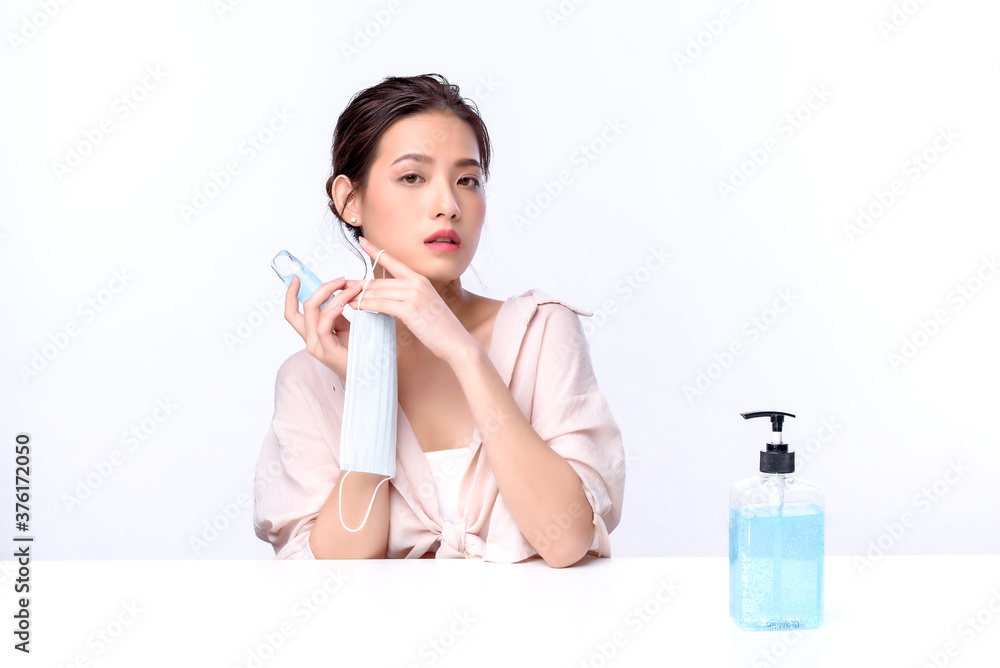 Portrait of beautiful charming young Asian woman posing with surgical mask or face mask to protect from corona virus and sitting with white table on white background, health care protection Covid-19 Portrait of beautiful charming young Asian woman posing with surgical mask or face mask to protect from corona virus and sitting with white table on white background, health care protection Covid-19