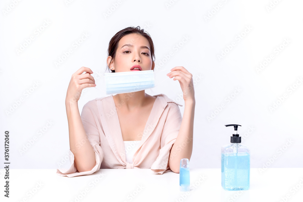 Portrait of beautiful charming young Asian woman posing with surgical mask or face mask to protect from corona virus and sitting with white table on white background, health care protection Covid-19 Portrait of beautiful charming young Asian woman posing with surgical mask or face mask to protect from corona virus and sitting with white table on white background, health care protection Covid-19