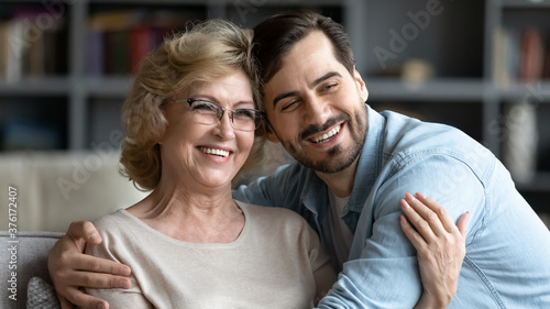 Overjoyed mature woman wearing glasses and adult son hugging, posing for photo, hugging, sitting on couch in living room, excited happy middle aged mother and young man embracing, two generations
