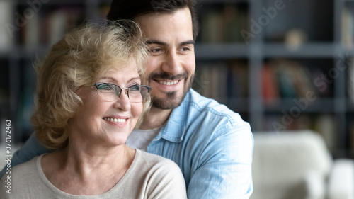 Close up smiling young man and mature mother wearing glasses dreaming together, happy dreamy middle aged woman and adult son hugging, looking to aside, dreaming about good future, standing at home