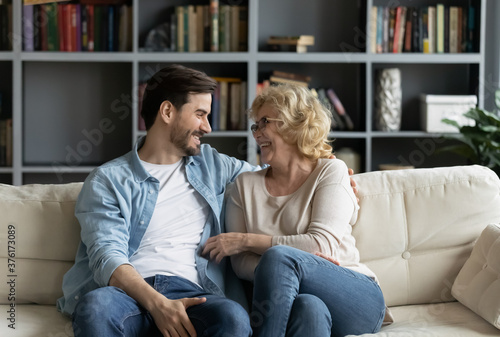 Happy mature woman wearing glasses and young man chatting, talking sitting on couch in living room, overjoyed elderly mother and adult son sharing news, spending weekend together at home