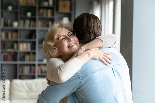 Close up smiling mature woman wearing glasses hugging adult son, standing in living room at home, family enjoying tender moment, beautiful happy middle aged woman embracing young man
