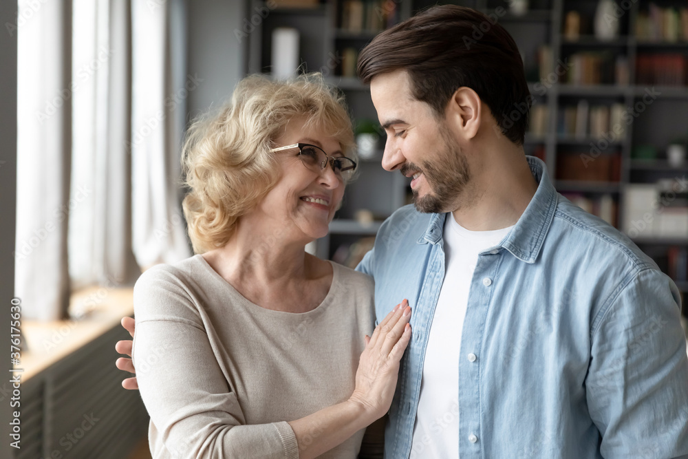 Close up smiling mature woman and adult son hugging, looking in eyes, family enjoying tender moment, standing in modern living room at home, happy elderly mother and young man cuddling