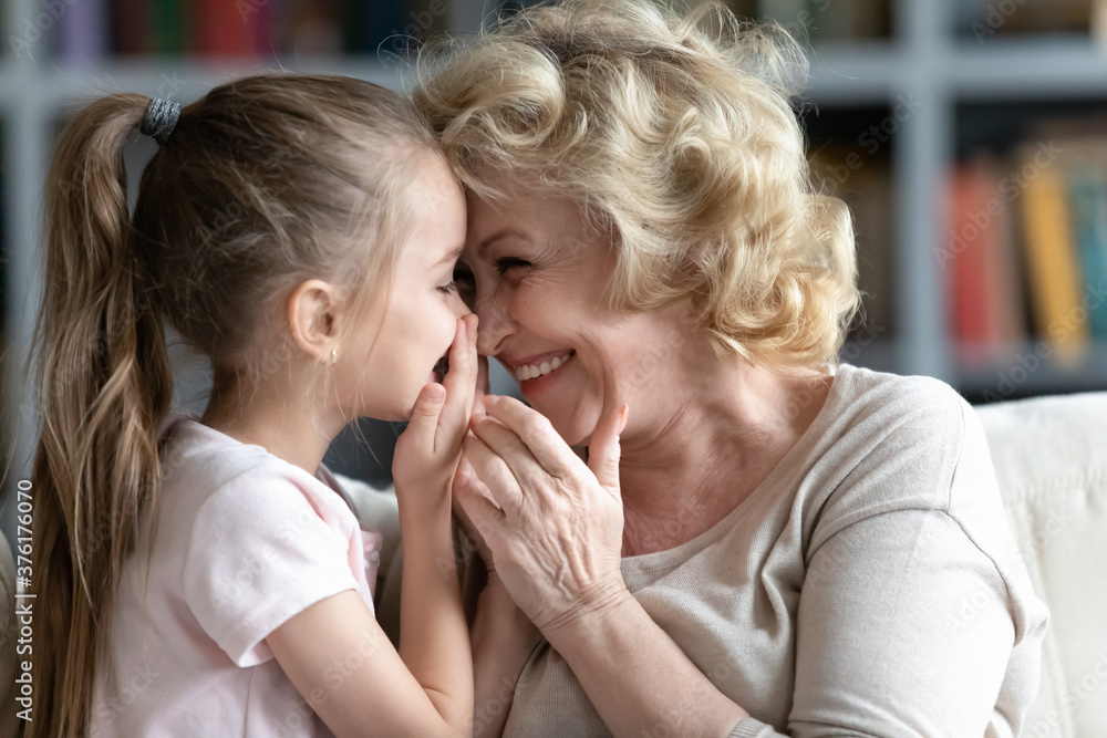 Close up smiling grandmother and little granddaughter talking, laughing, pretty preschool girl sharing secrets with happy grandma, whispering, having fun, sitting on cozy couch at home Close up smiling grandmother and little granddaughter talking, laughing, pretty preschool girl sharing secrets with happy grandma, whispering, having fun, sitting on cozy couch at home