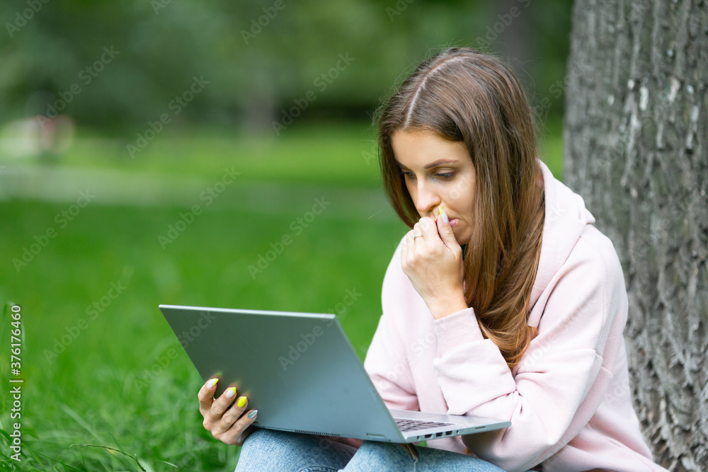 Pensive student girl having a video call, while sitting on the grass in campus park. Pensive student girl having a video call, while sitting on the grass in campus park.