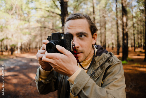 Young male photographer focussing while taking pictures with film camera in bright forest.