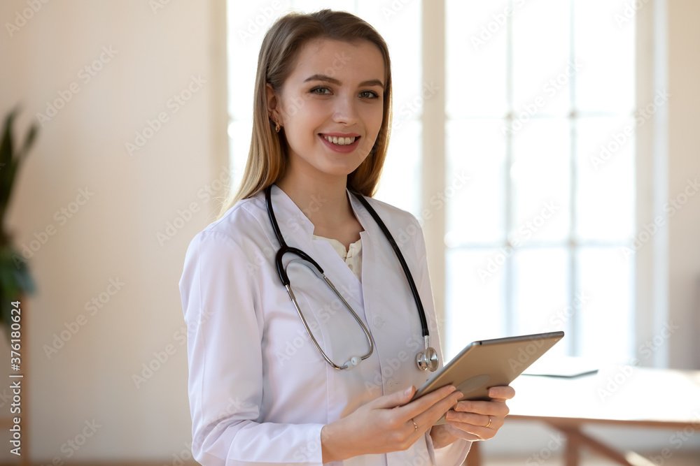 Head shot portrait smiling young woman doctor wearing white uniform with stethoscope with computer tablet, standing in modern office in hospital, confident therapist physician gp working online Head shot portrait smiling young woman doctor wearing white uniform with stethoscope with computer tablet, standing in modern office in hospital, confident therapist physician gp working online