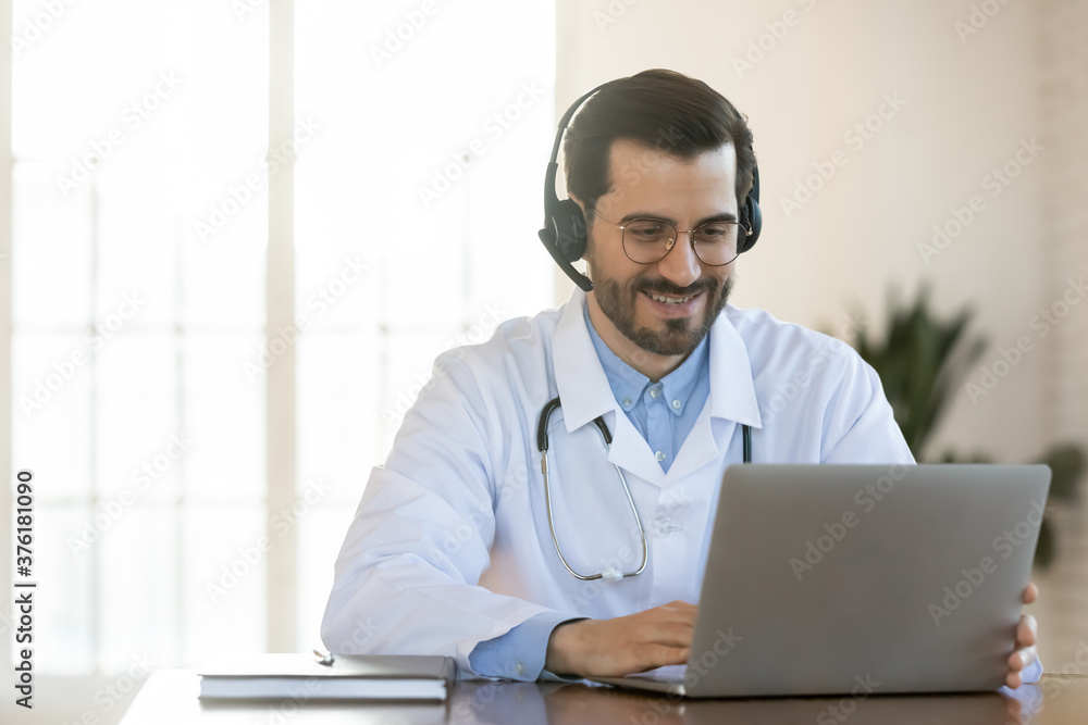 Smiling young doctor in headset consulting patient online, using laptop, looking at screen, sitting at desk in office, therapist gp wearing white uniform making video call, telemedicine concept Smiling young doctor in headset consulting patient online, using laptop, looking at screen, sitting at desk in office, therapist gp wearing white uniform making video call, telemedicine concept