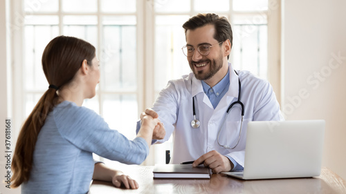 Smiling young doctor wearing white uniform with stethoscope shaking female patient hand at meeting, therapist gp wearing glasses greeting woman at medical appointment in office, sitting at table