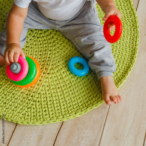 Little child boy plaing with toy pyramid on knitted carpeet on the wooden floor indoor. Knitted style in the interior. Warm and cozy. Playful children