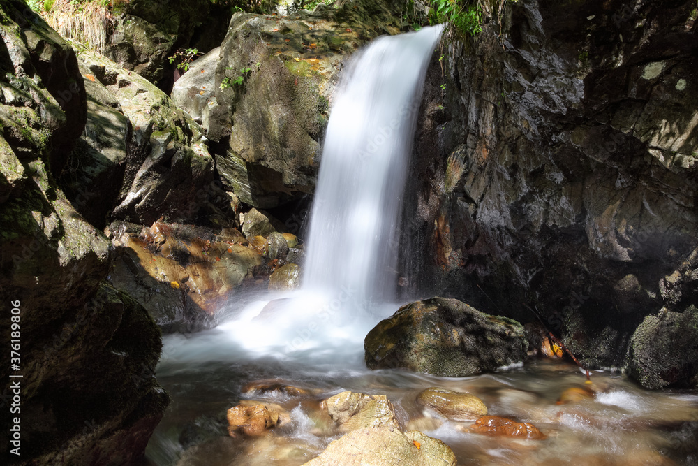 Fototapeta premium Mountain waterfall flows among green forest and runs down the beautiful gray stones. Amazing summertime wallpaper background.