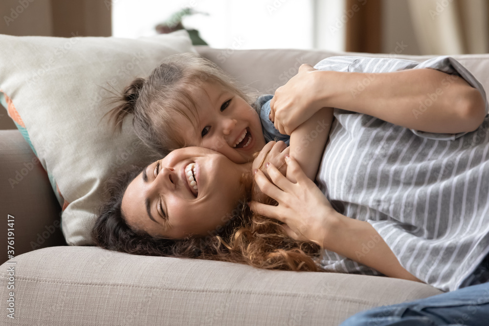 Close up overjoyed mother enjoy sweet tender cuddles of small cheerful daughter lying on sofa laughing having fun at home. Happy motherhood, priceless time together, active games with children concept Close up overjoyed mother enjoy sweet tender cuddles of small cheerful daughter lying on sofa laughing having fun at home. Happy motherhood, priceless time together, active games with children concept
